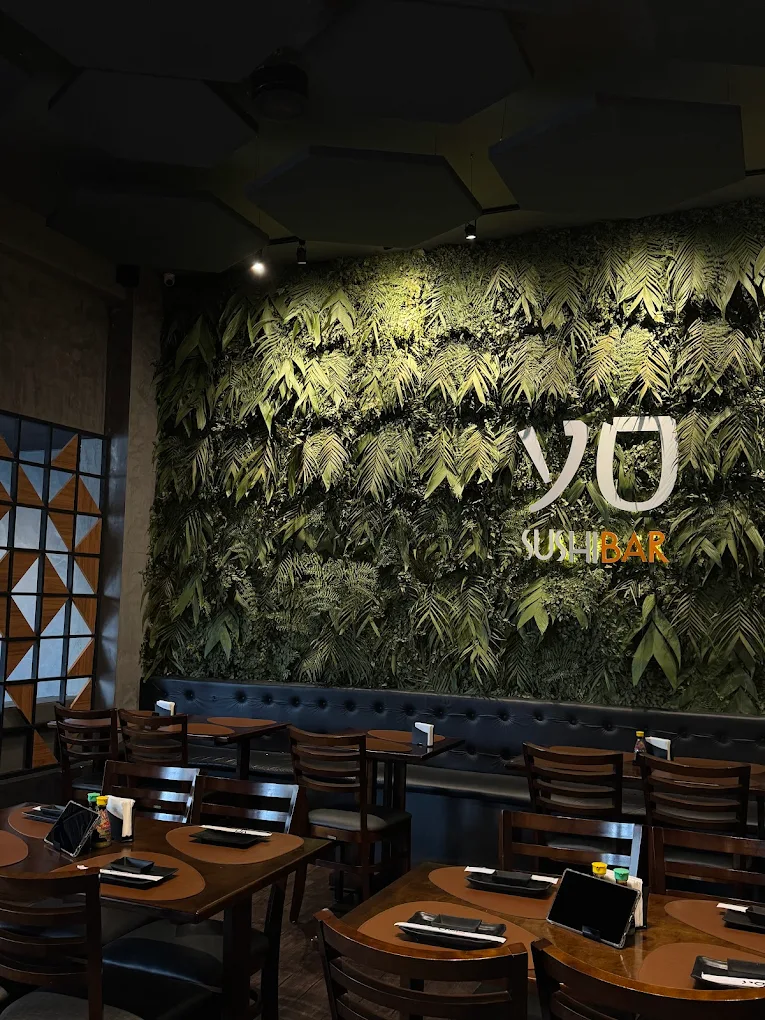 Interior of a sushi bar with wooden tables, dark chairs, and a large green plant wall featuring the words 'YO SUSHI BAR'.